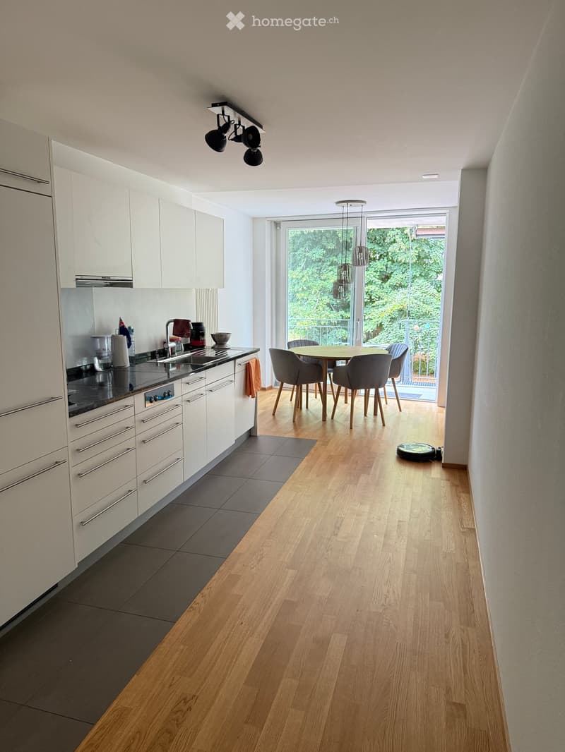 Modern kitchen with white cabinets, dark countertops, and a dining area with a round table and chairs. The space features wooden flooring and large windows with a view of greenery.