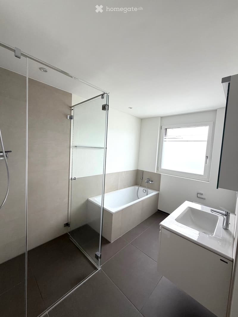 Modern bathroom with a bathtub, sink, glass shower enclosure, and a window providing natural light.