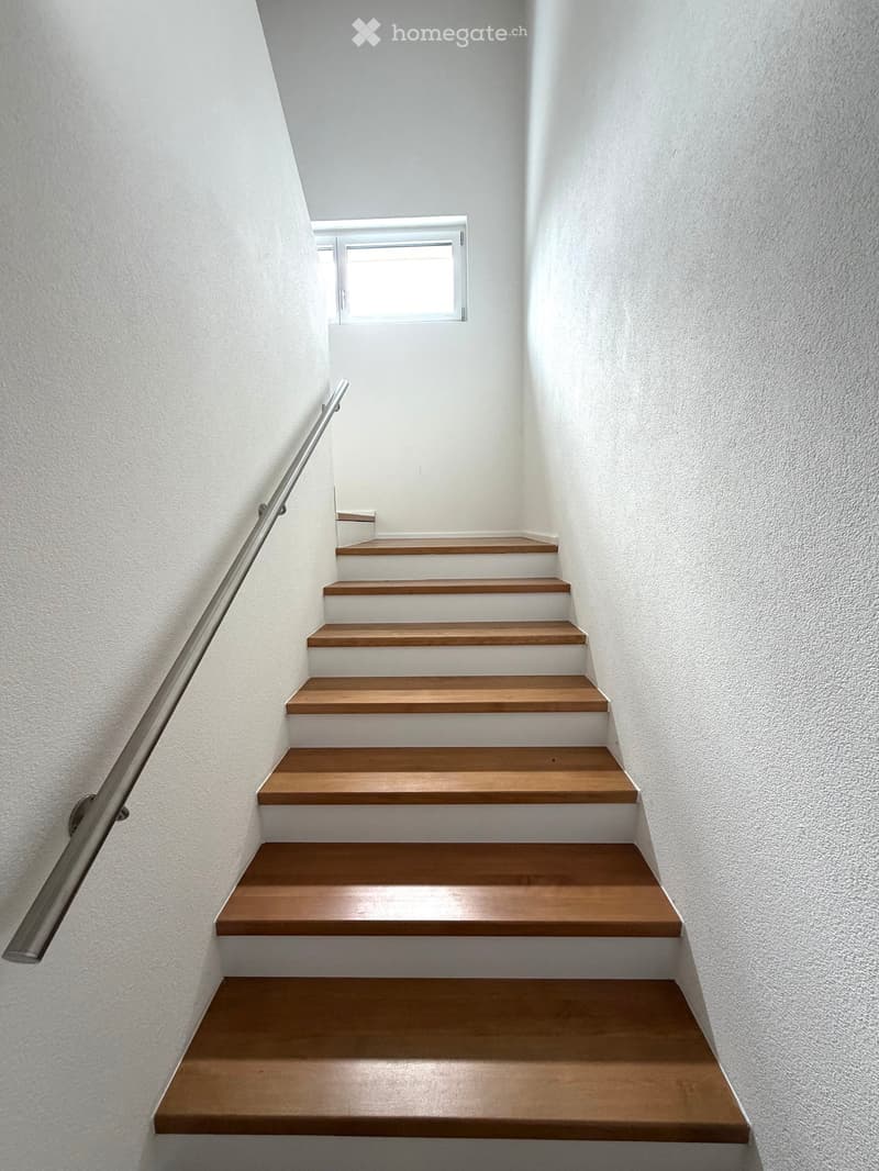 Modern staircase with wooden steps and white risers, featuring a stainless steel handrail and a small window at the top.