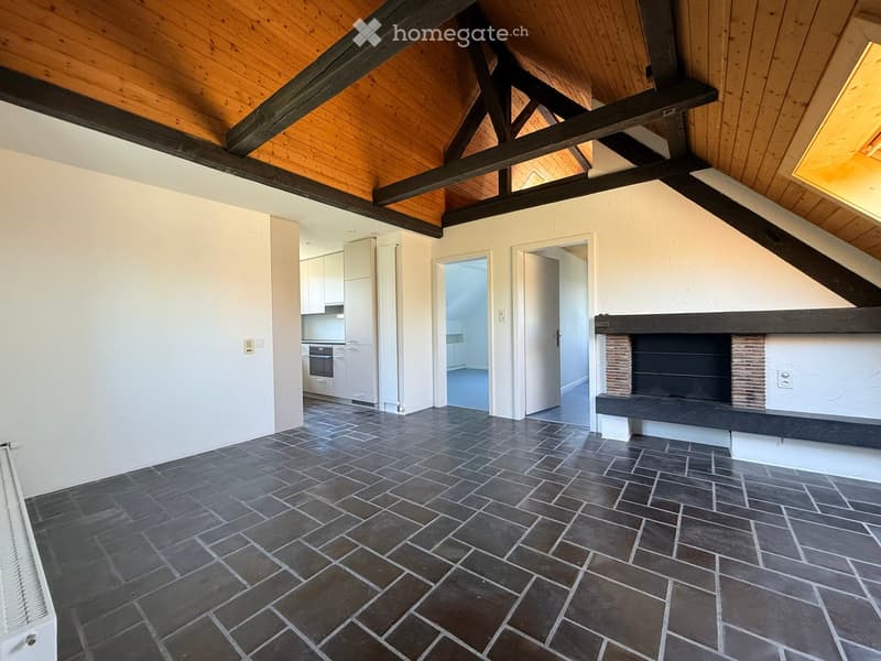 Loft-style room with exposed wooden beams, dark tiled floor, fireplace, radiator, and open kitchen area visible in the background.