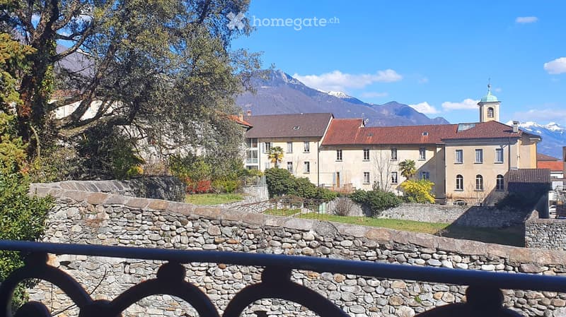 A scenic view of a town with traditional buildings, red-tiled roofs, and a stone wall in the foreground, with mountains in the background.