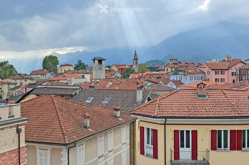 A panoramic view of a historic town with red-tiled roofs, multiple buildings, and a church tower, set against a backdrop of mountains and a cloudy sky.