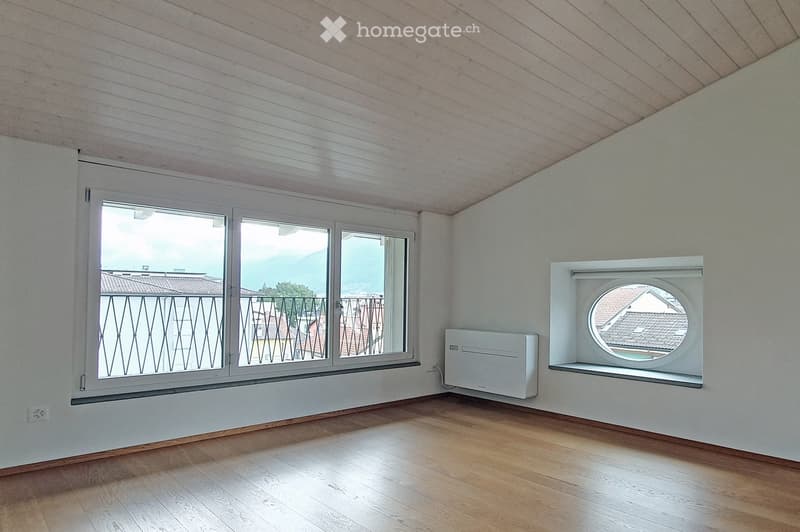 Empty room with wooden flooring, white walls, and large windows offering a view of a balcony and surrounding buildings.