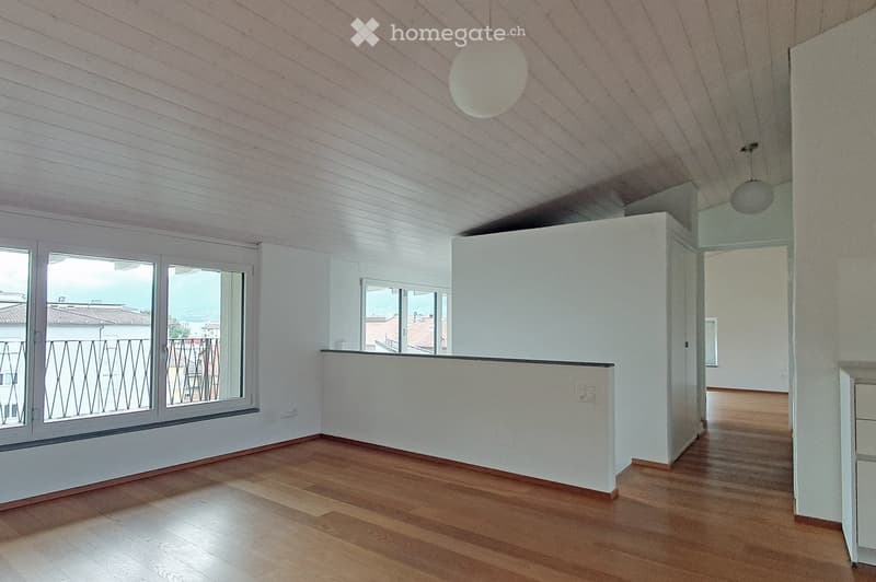 Empty room with wooden flooring, white walls, large windows with metal railings, and a slanted ceiling with pendant lights.