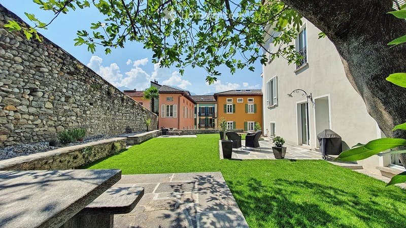 Well-maintained garden with green lawn, stone benches, outdoor seating area with chairs and table, potted plants, and surrounding buildings.