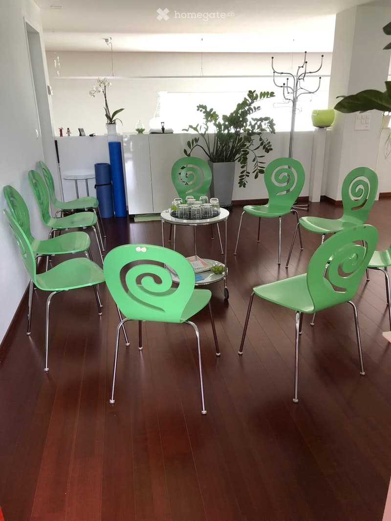 A modern, well-lit room with wooden flooring, several green chairs with spiral backrests, a small round table with cups, and a coat rack near the entrance.