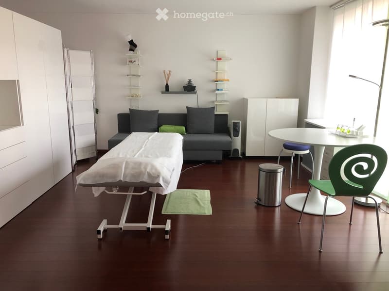 Modern living room with dark wooden flooring, white walls, and large windows. Features a grey sofa with dark cushions, a white round table with a green chair, a white cabinet, a shelf with decorative items, a floor lamp, and a massage table covered with a