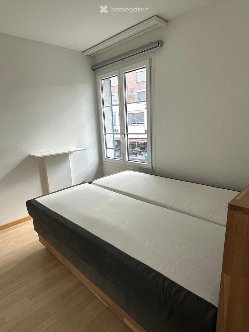 A small bedroom with a single bed, wooden flooring, and a window with blinds. The bed has a white mattress and dark grey bedding.