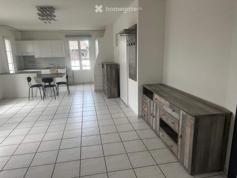 Open-plan living and dining area with tiled flooring, white walls, and modern furniture including a wooden cabinet, a table with chairs, and a small kitchen area with cabinets and a sink.