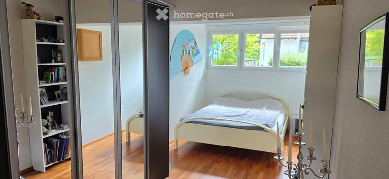 Cozy bedroom with wooden flooring, a single bed with white bedding, a bookshelf filled with books and decorative items, and a large mirror on the wall.
