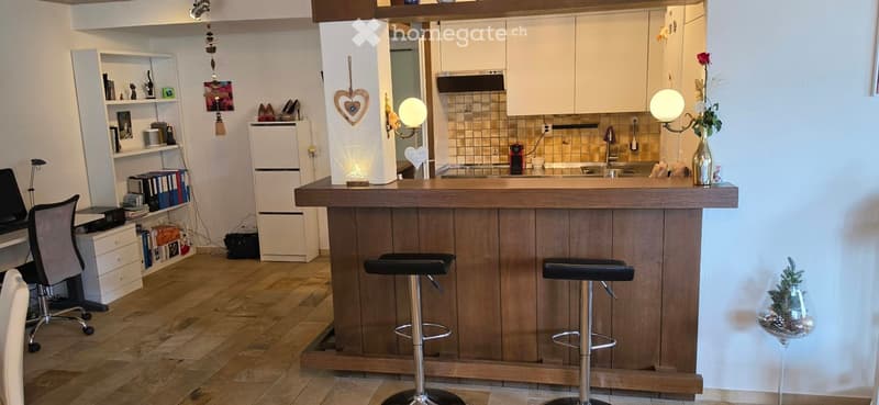 Modern kitchen with a wooden bar counter, two black stools, white cabinets, a refrigerator, and a bookshelf with books and decorative items.