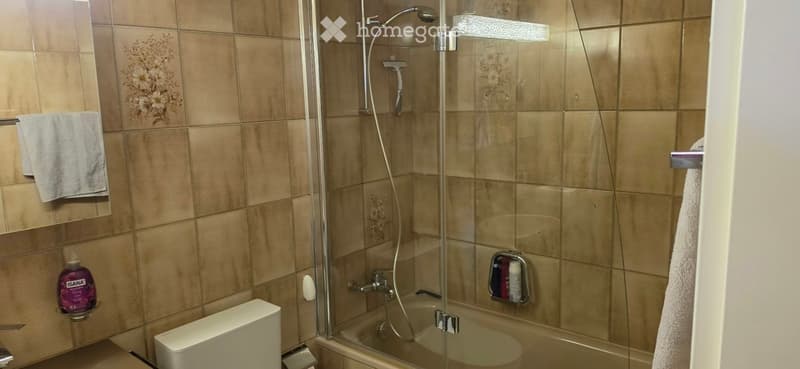 Modern bathroom with beige tiles, a bathtub with a glass shower enclosure, a toilet, a towel, and a mirror.