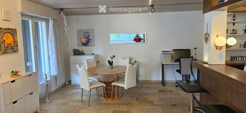 Well-lit room with a round dining table surrounded by white chairs, a desk with a computer, a bar stool, and decorative shelves.