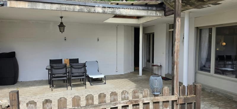 Outdoor patio with a small table and chairs, a wicker basket, a decorative vase, and a small stool, connected to a house with large windows.