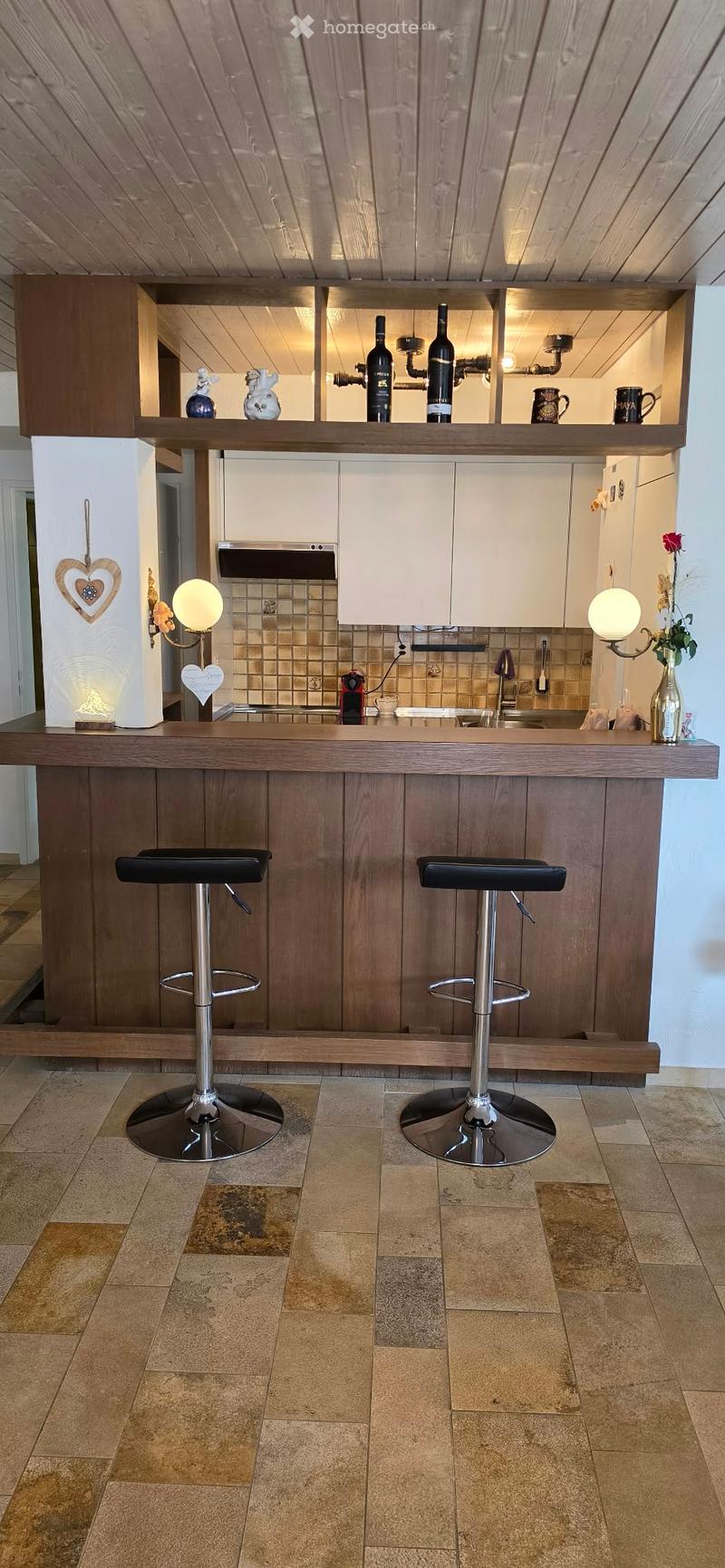 Modern kitchen with a wooden bar counter, two black stools, white cabinets, and a tiled backsplash. Shelves hold bottles, cups, and decorative items. A vase with flowers and a lamp are on the counter.