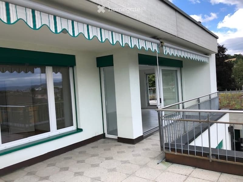 Balcony with striped awning, metal railing, and tiled floor, leading to a glass door of the building.