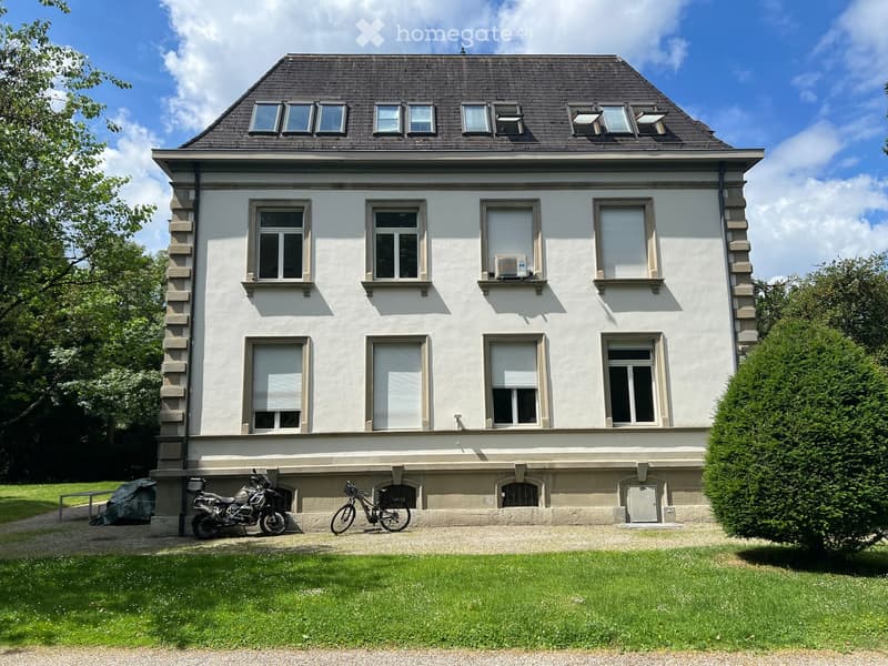 Two-story white house with multiple windows, a dark roof, and a well-maintained lawn in front. There is a motorcycle and a bicycle parked near the entrance.