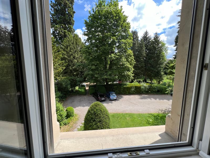View from a balcony showing a well-maintained garden with trees, shrubs, a gravel driveway, and two parked cars.
