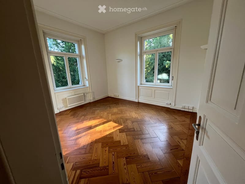 Empty room with wooden herringbone flooring, white walls, and large windows with white frames. Natural light streams in, illuminating the space.