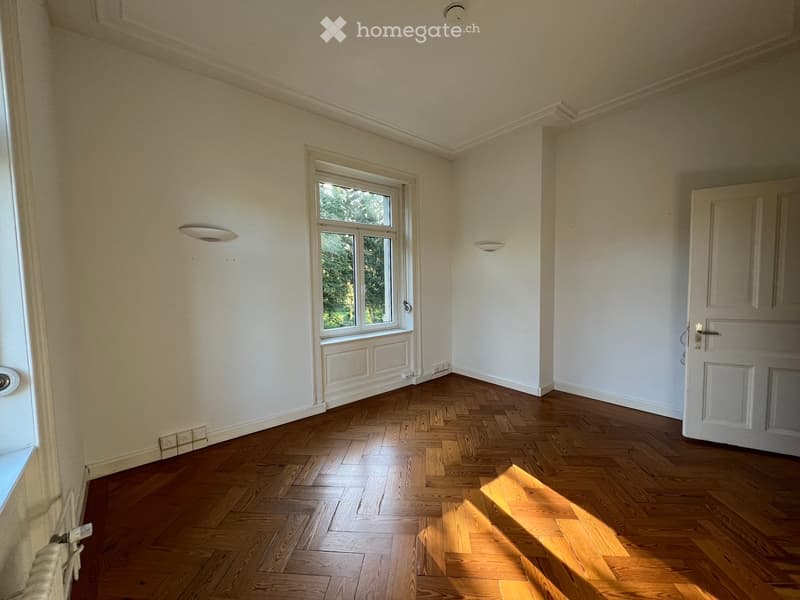Empty room with wooden herringbone flooring, white walls, and a large window letting in natural light. The room features built-in wall panels and recessed lighting.