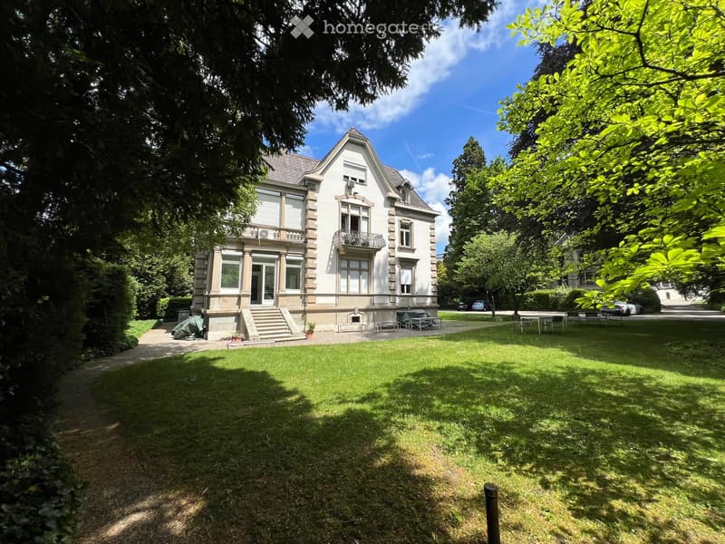 A large, elegant two-story house with white exterior and multiple windows, surrounded by a well-maintained lawn and trees. Features include a front staircase, a small porch area, and outdoor seating.