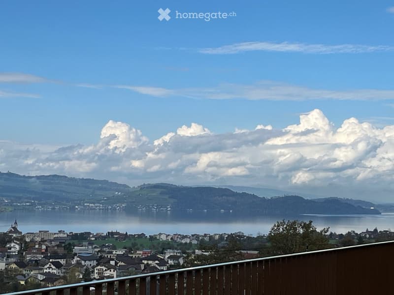 Panoramic view from a balcony overlooking a town, lake, and distant hills under a partly cloudy sky.