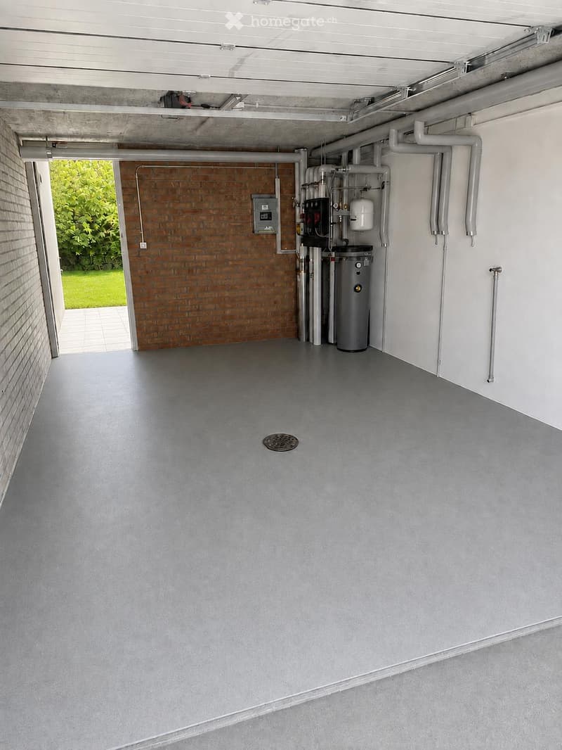 Empty utility room with grey concrete floor, white walls, and a brick wall section. Visible pipes, a water heater, and an electrical panel are mounted on the walls.