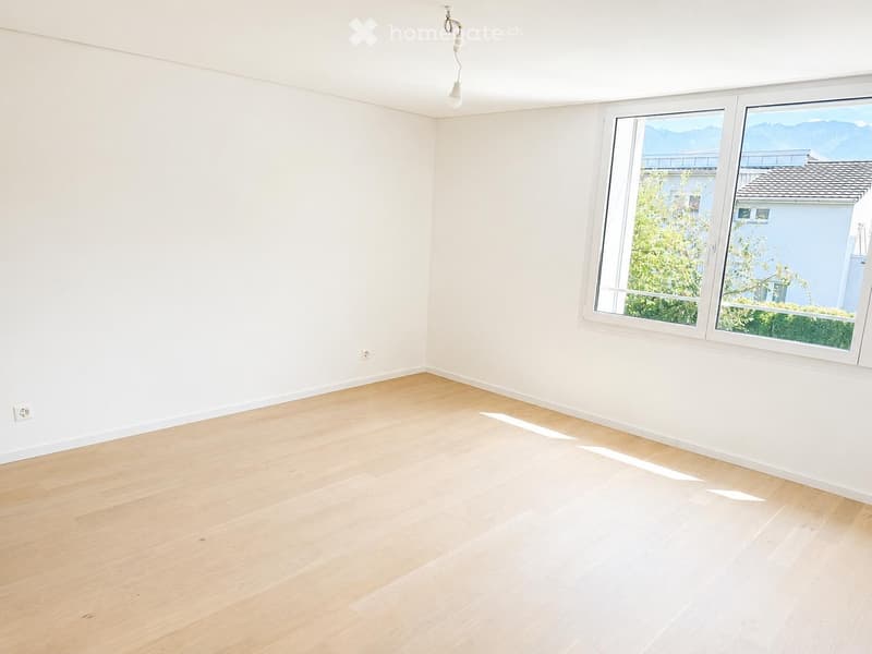 Empty room with light wooden flooring and white walls, featuring large windows with a view of greenery and mountains.