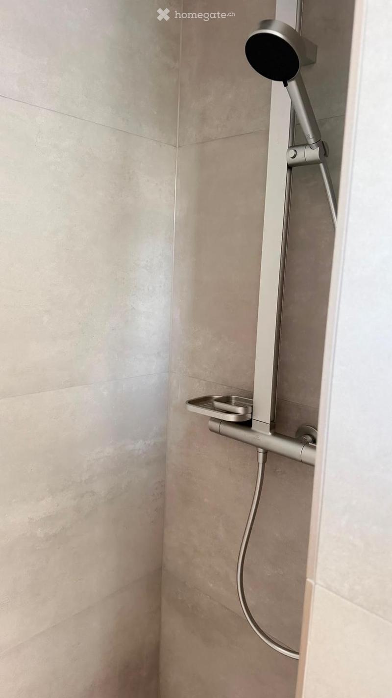 Modern showerhead with handheld attachment mounted on beige tiled wall.