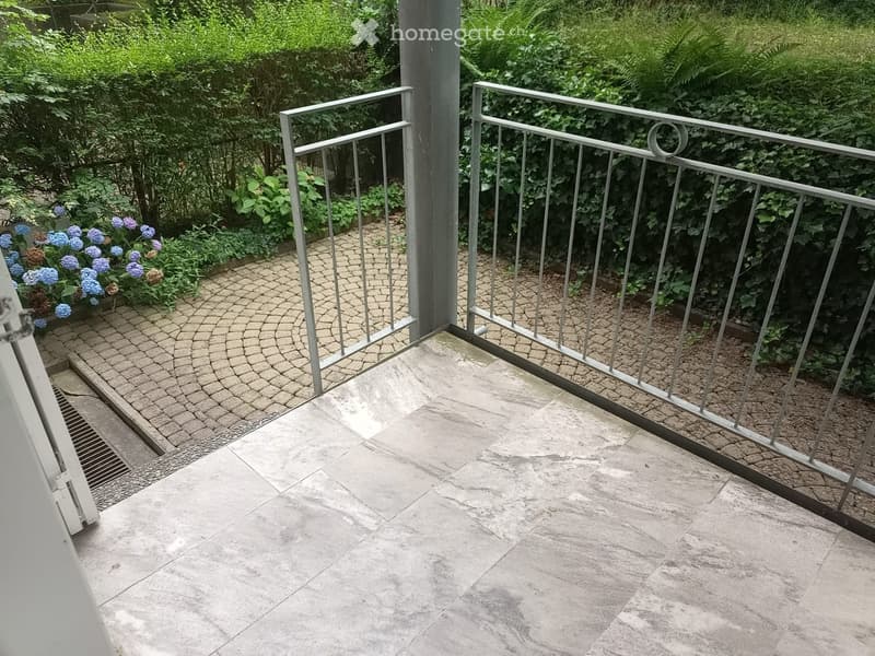 Balcony with stone tiles, metal railing, and a view of a garden with flowers and shrubs.