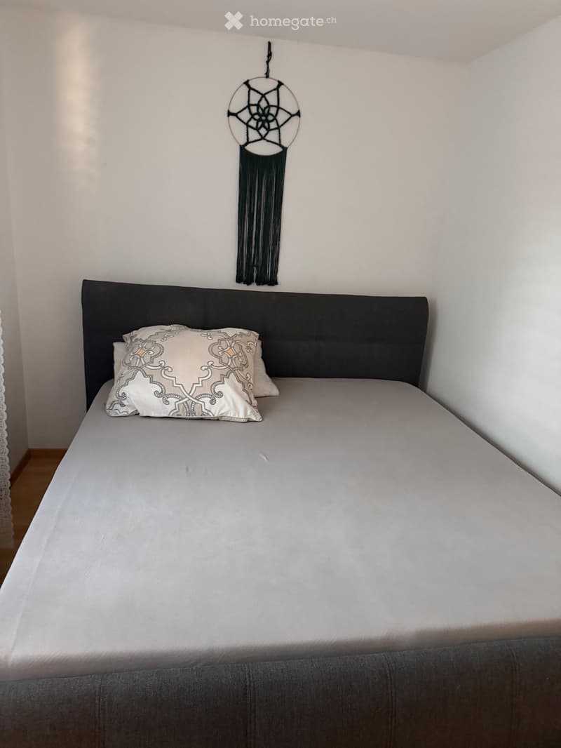 A neatly made bed with a dark headboard, white bedding, and decorative pillows, complemented by a wall hanging with a circular design and long tassels.