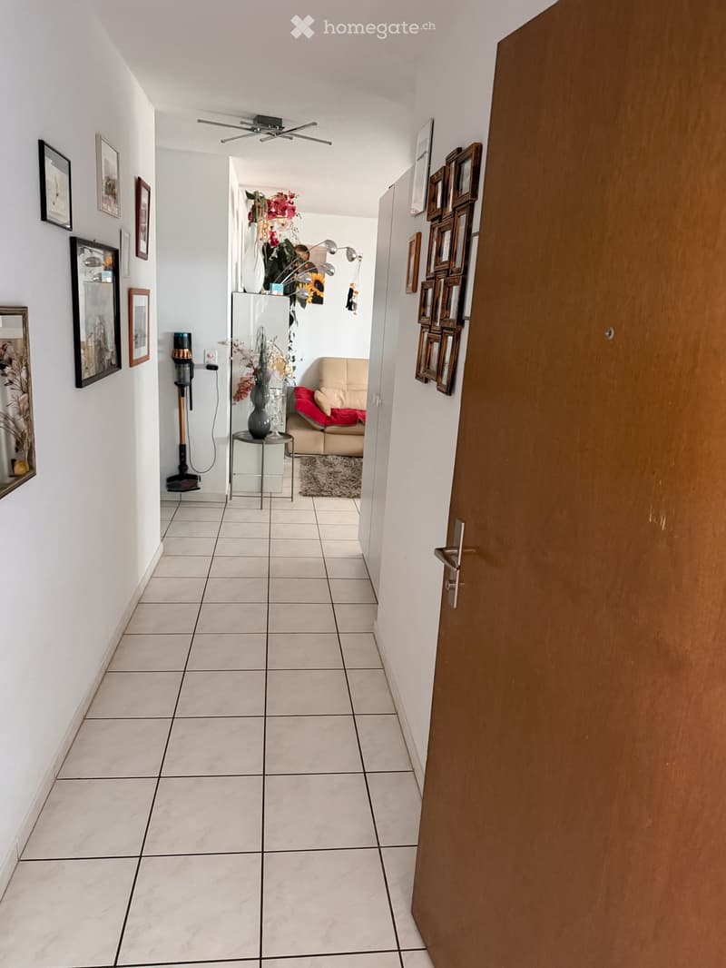 Clean hallway with tiled floor, white walls, framed pictures, and a small table with vase and flowers leading to a living room area with a sofa and rug.