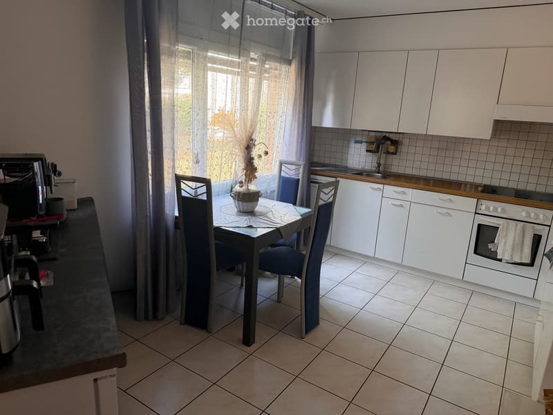 Modern kitchen with white cabinets, tiled floor, and a dining area featuring a table and chairs. A vase with dried flowers is placed on the table.
