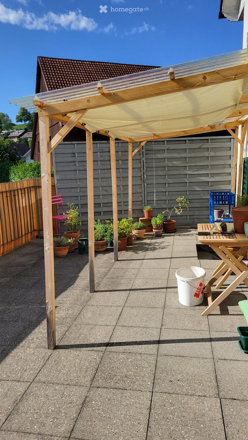 Outdoor patio with a wooden pergola, potted plants, a small table, and chairs.