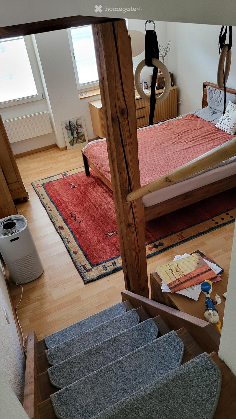 Cozy bedroom with a wooden bed, nightstand, books, a small fan, and a gymnastics ring hanging from the ceiling. The room features wooden flooring and a staircase leading to another level.