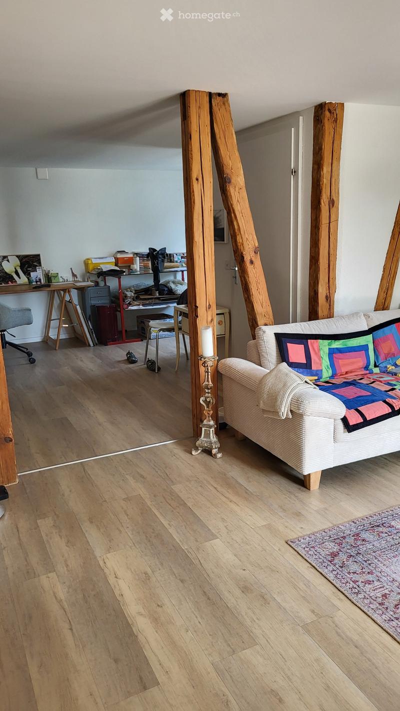 A cozy living space with wooden floors, a white sofa with colorful pillows, a small table with a candle holder, a desk with office supplies, and a rug on the floor.