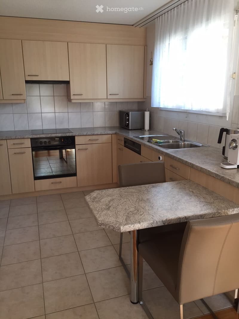 Modern kitchen with light wood cabinets, granite countertops, stainless steel appliances, and a small dining area with a chair and table.