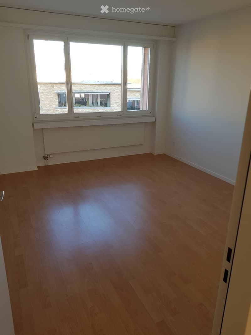 Empty room with wooden flooring, white walls, and large windows with white frames, offering a view of a brick building outside.