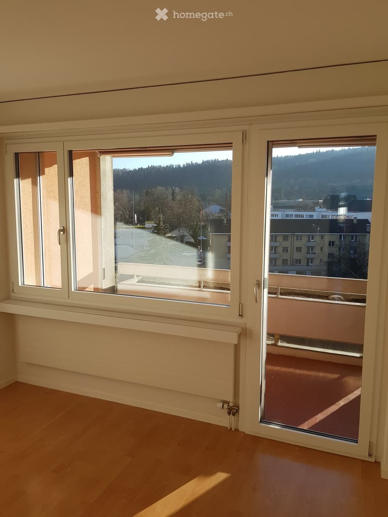 Empty room with wooden flooring, large windows, radiator, and a balcony with a view of the city and surrounding landscape.