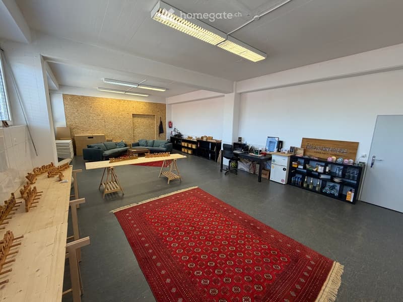 The room features a sofa, coffee table, chairs, a large wooden table, shelves, a refrigerator, and a desk with a chair. The space is well-lit with overhead lighting and includes a red patterned rug on the floor.