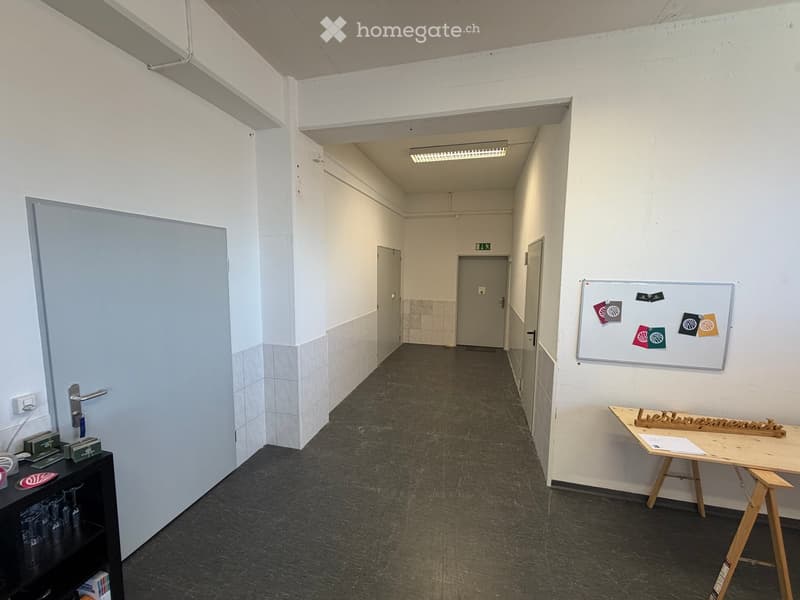 Empty hallway with white walls, grey tiled lower sections, and a black floor. Features a whiteboard with colorful magnets, a small wooden table with papers, and a black shelf with glassware and books.