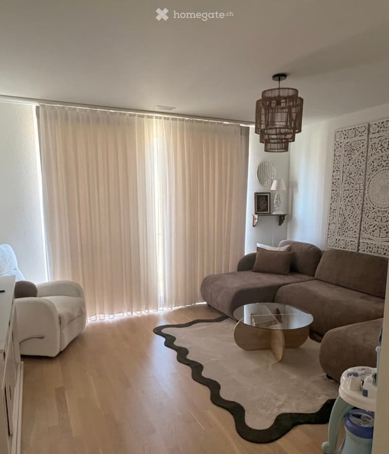 Modern living room with a large window covered by beige curtains, a brown sectional sofa, a glass coffee table, a white armchair, and a decorative wall piece.