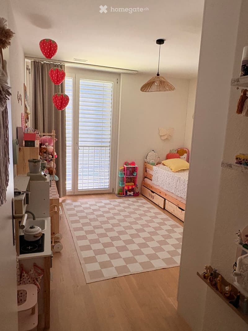 A well-lit bedroom with a bed, a small kitchen area, and various toys and decorations. The room features a checkered rug, a hanging lamp, and a window with blinds leading to a balcony.