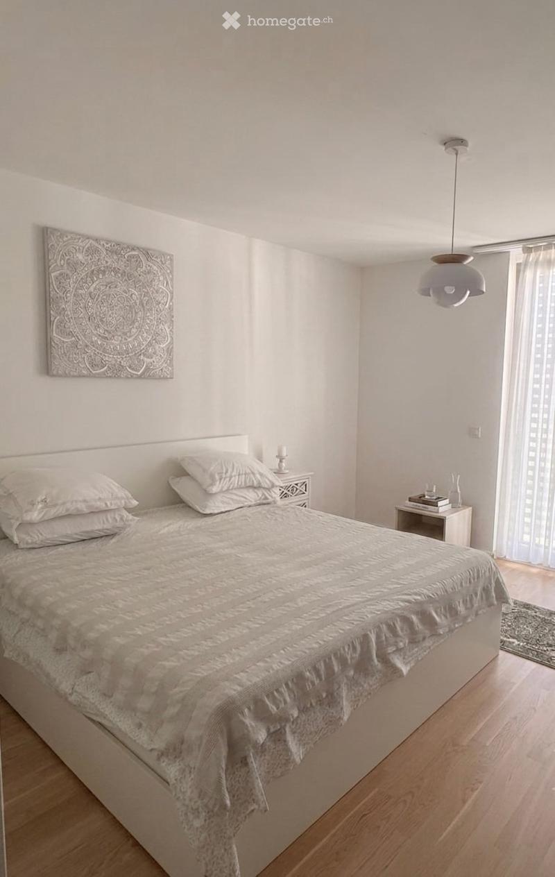 Modern bedroom with a large bed, white bedding, nightstand, lamp, decorative wall art, and a vase on a small table near the window.