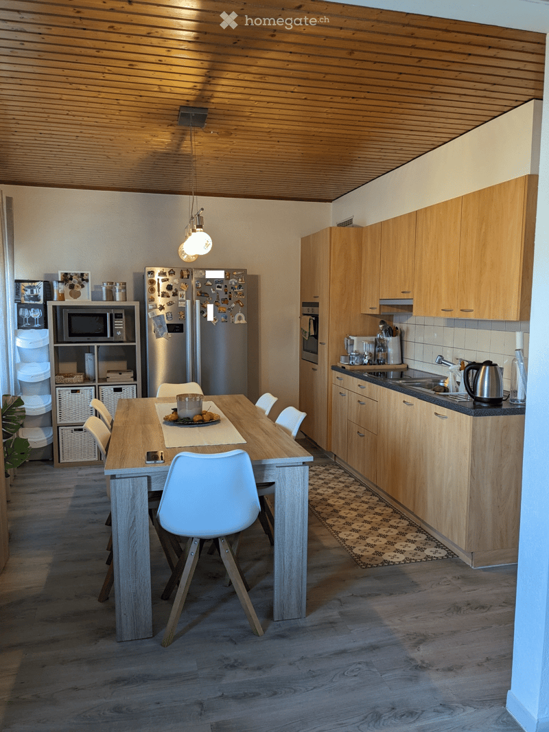 Modern kitchen with wooden cabinets, stainless steel appliances, a dining table with chairs, and a pendant light fixture.