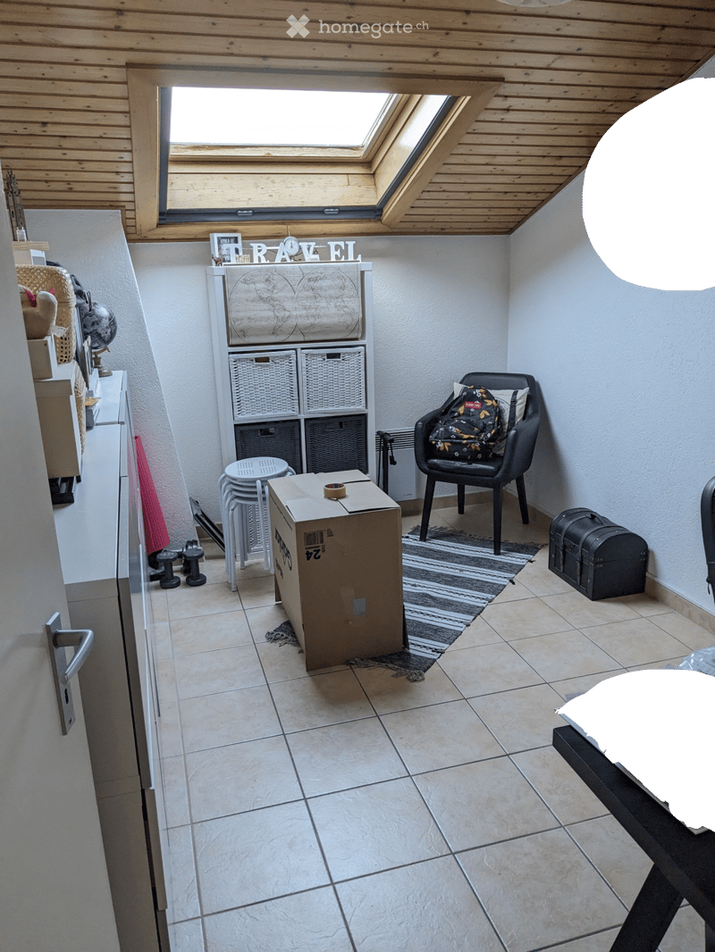 A small, furnished room with a skylight, tiled floor, and various items including a chair with a backpack, a storage unit with baskets, a globe, a lamp, and a small table.