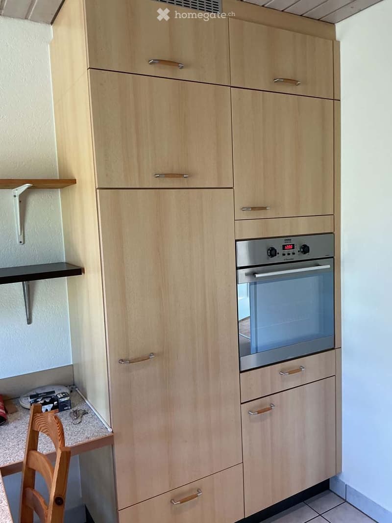 Modern kitchen with light wood cabinets, stainless steel oven, and a small wooden chair near the counter.