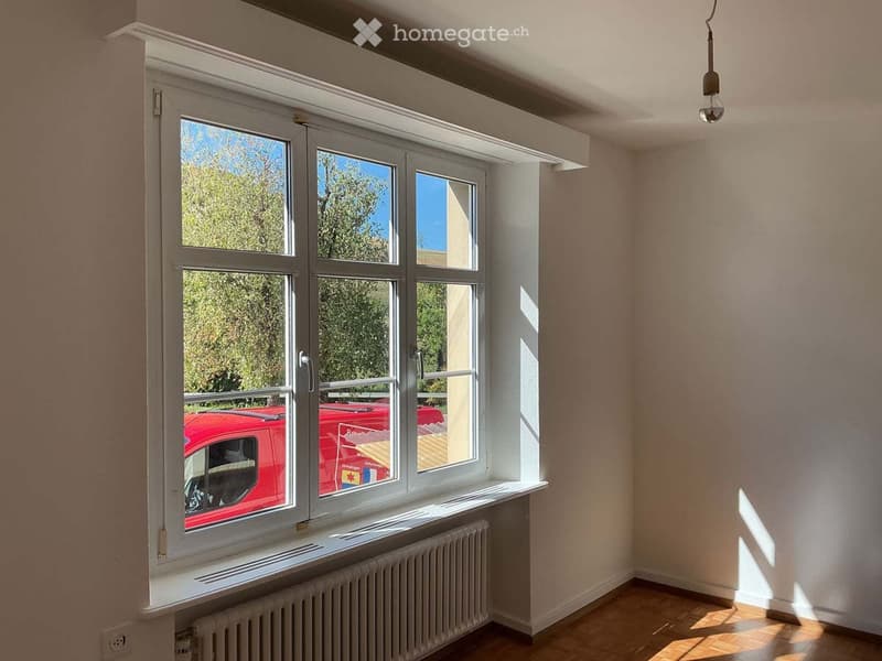 Empty room with large windows, wooden flooring, radiator, and a hanging light fixture. No furniture or equipment present.