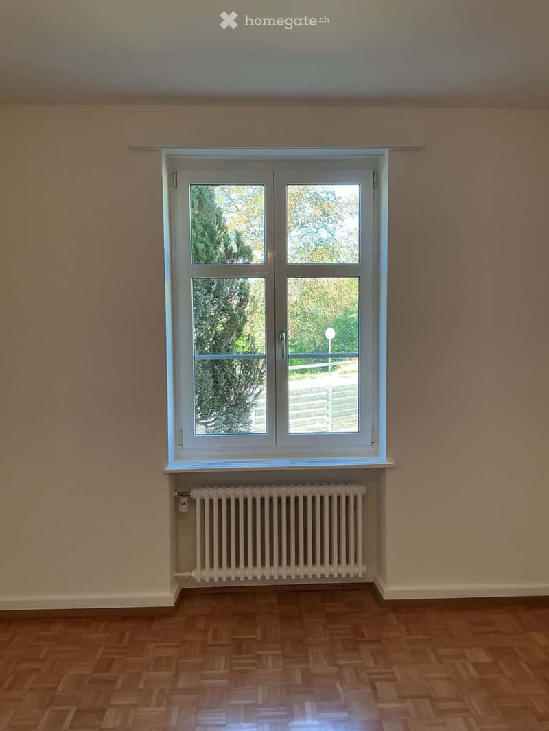 Empty room with wooden flooring, white radiator, and large window showing outdoor view.