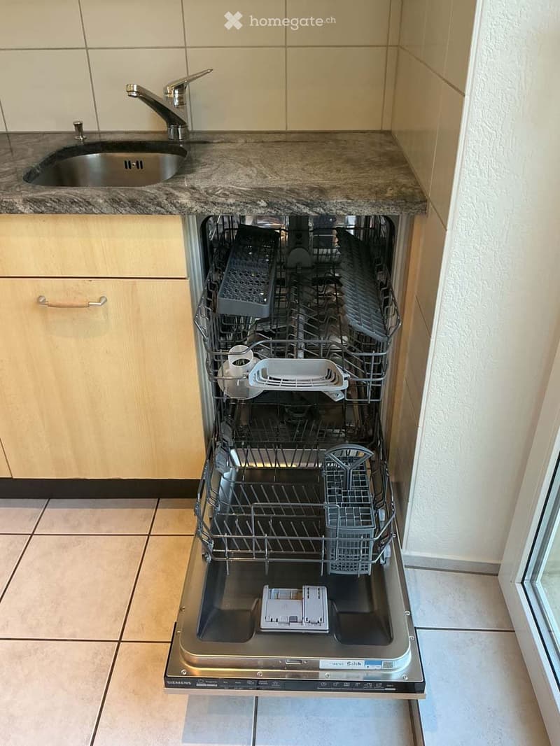 Open stainless steel dishwasher with racks and dishes inside, placed under a granite countertop with a sink and faucet.